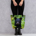 Tote bag with black bear cub photo print, held by person against gray wall background