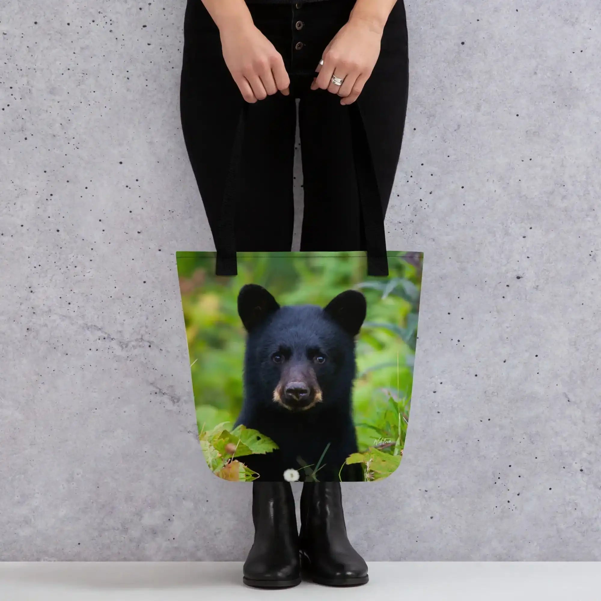Tote bag with black bear cub photo print, held by person against gray wall background