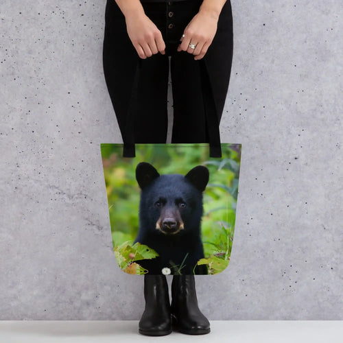 Tote bag with black bear cub photo print, held by person against gray wall background
