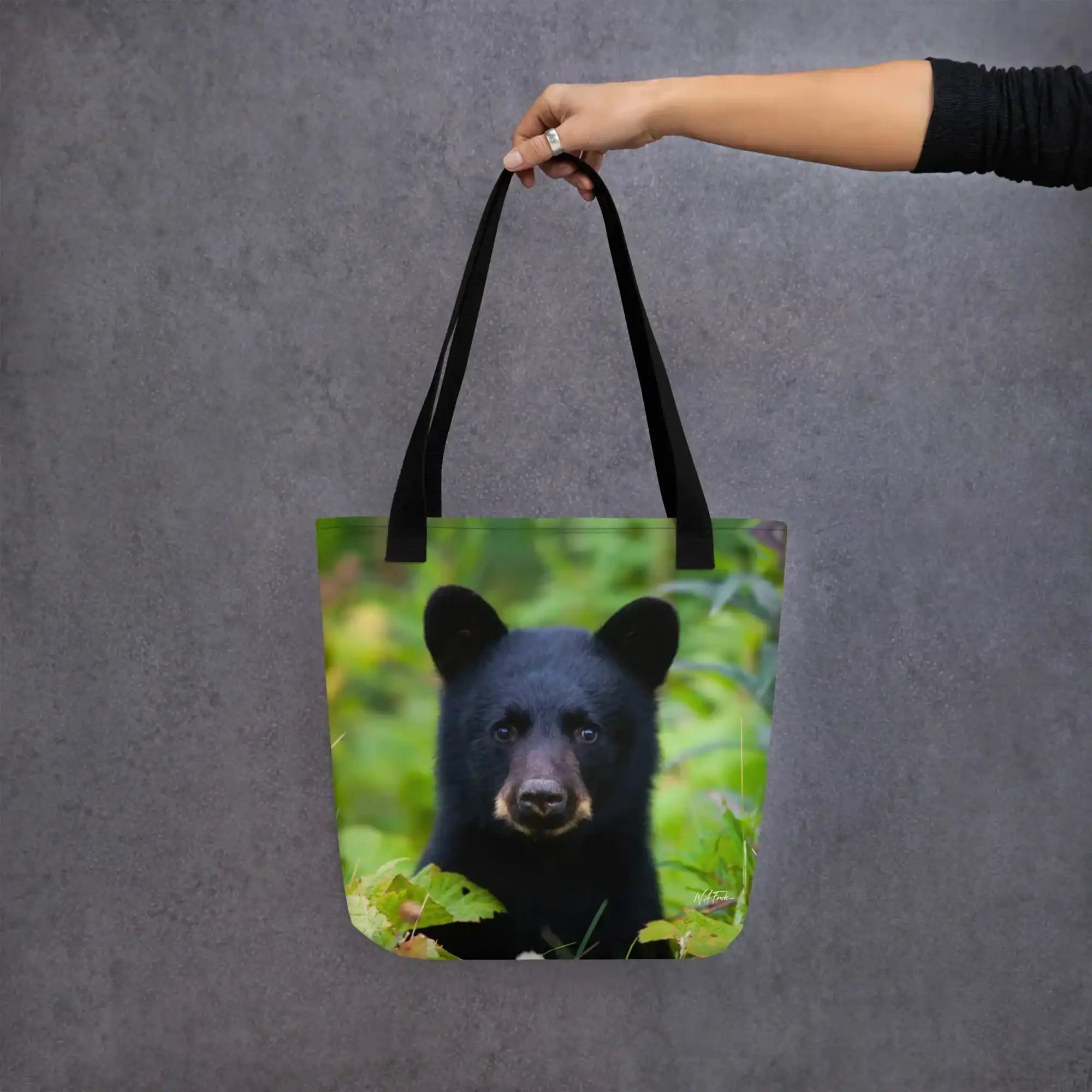 Black tote bag with a photo of a bear cub in green foliage, held by a hand against gray background