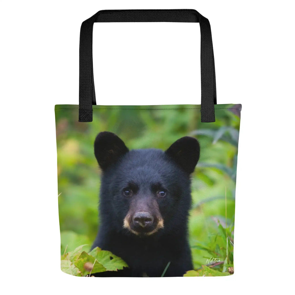 Tote bag with black bear cub photo and black handles, set against a green foliage background.